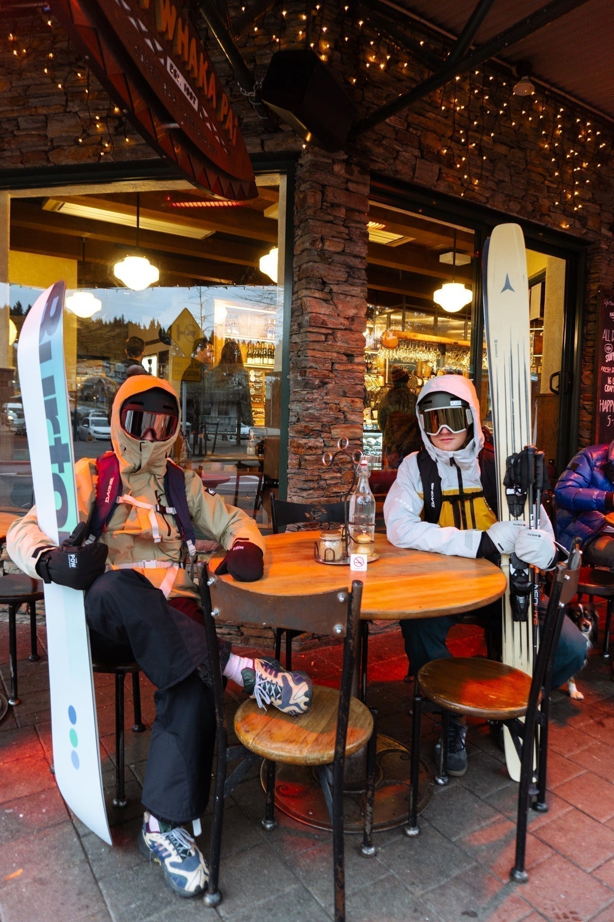 Two riders in ski gear and goggles at an outdoor table under the string lights at Kai Whakapai, Wānaka
